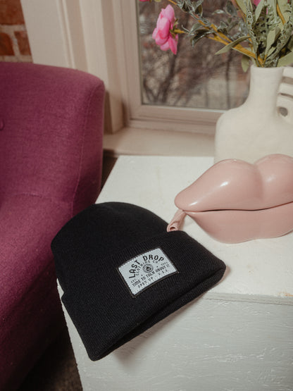 A black beanie with a woven embroidered patch sits on a table next to a pink vase.